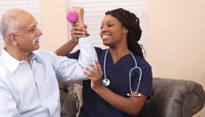 Senior adult, male patient does arm exercises with his African descent home healthcare nurse or physical therapist in nursing home or home setting.  Patient uses dumbbell.